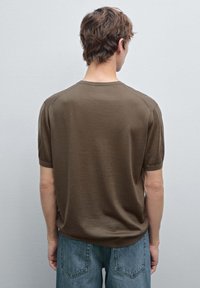 Hnědý pletený svetr s krátkým rukávem a kulatým výstřihem. Měkká textura a volný střih. Nosený s světle modrými džíny, viděno ze zadu.