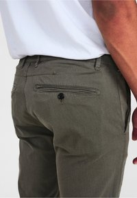 Närbild av en person som bär texturerade olivgröna byxor med en knäppt bakficka och en instoppad vit skjorta.