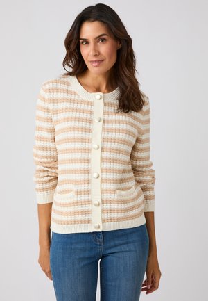 Femme portant un cardigan rayé beige et blanc boutonné avec des poches avant et un jean bleu, debout devant un fond uni.