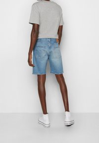 Person som bär ljusblå denimshorts, grå t-shirt med kort ärm och vita högklackade sneakers, stående mot en enfärgad ljus bakgrund.