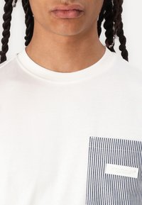 Tricou alb din bumbac cu guler rotund, având un buzunar în dungi bleumarin. Buzunarul are o etichetă albă cu text. Textură moale și netedă.