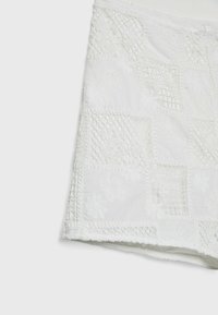 Jupe en tissu blanc avec broderie patchwork comprenant des motifs floraux et en treillis sur un fond blanc uni.