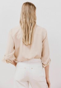 Camicia beige con bottoni e maniche arrotolate indossata con jeans bianchi a vita alta con dettagli dorati e tasche posteriori. Capelli che cadono oltre le spalle.