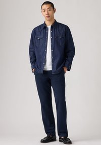 Homme portant une chemise en jean bleu foncé, un t-shirt blanc, un pantalon marine et des chaussures noires, debout les mains dans les poches devant un fond uni.