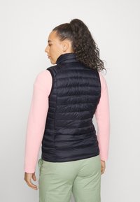 Marineblå pufferväst med hög krage, quiltad textur och figursydd siluett, bärs över en rosa långärmad skjorta och gröna byxor.