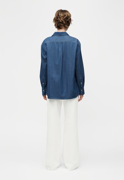Άτομο που στέκεται με την πλάτη προς τα πίσω, φορώντας μπλε τζιν πουκάμισο με μακριά μανίκια και λευκό φαρδύ παντελόνι, μπροστά σε απλό φόντο.