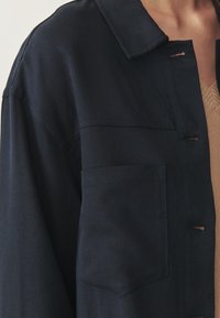 Tume sinine jakk koos kraega, eesmine tasku ja nööbiga detailid. Valmistatud siledast kangast, millel on peen tekstuur.