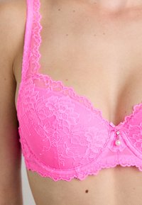 Rosa Spitzen-BH mit Blumenmuster, weichem Material, verstellbaren Trägern und einem dekorativen Perlenakzent in der Mitte.