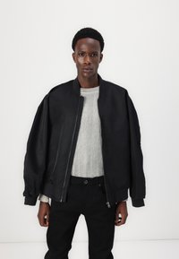 Mladý muž s krátkymi kučeravými vlasmi, oblečený v čiernej prelage nadmernej veľkosti, pod ktorou má svetlošedý rebrovaný sveter a čierne nohavice, stojí proti bielej pozadí.