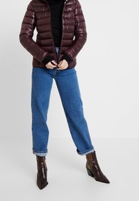 Burgundy puffer jacket with horizontal quilting, black cuffs, and a fitted design, paired with blue loose-fit jeans and patterned brown ankle boots.