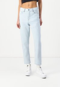 Personne portant un jean bleu clair taille haute à jambes droites et des baskets blanches montantes, debout sur un fond blanc uni.