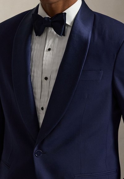Férfi sötétkék selyem shaalgalléros szmokingzakóban, fehér redős ingben, fekete patent gombokkal és hozzá illő sötétkék csokornyakkendőben.