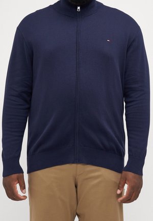 Homme portant un pull zippé bleu marine et un pantalon beige sur fond blanc, visible du cou aux hanches.