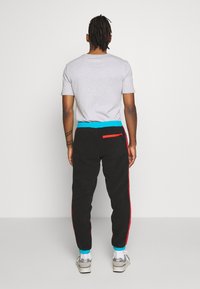 Pantalones de chándal negros con rayas laterales rojas, que presentan un cordón azul en la cintura y un bolsillo con cremallera en la parte derecha. Está visible una camiseta gris en la parte superior.
