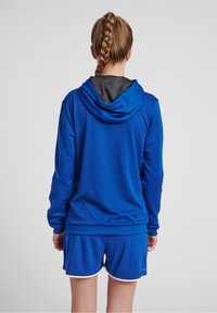 Felpa sportiva blu con rifiniture nere, tessuto strutturato e maniche lunghe, abbinata a pantaloni corti blu coordinati con dettagli bianchi.