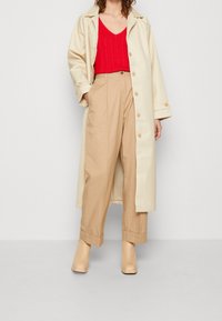 Krémový dlouhý kabát, červený pletený top, béžové široké kalhoty a tělové platformové podpatky. Hladké textury s knoflíky na kabátu a pleteným vzorem na topu.