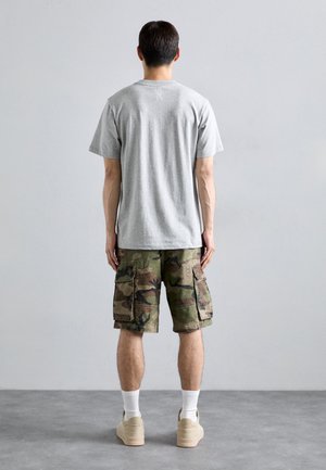 T-shirt grigio in cotone con maniche corte, abbinato a pantaloni cargo mimetici verdi con diverse tasche. Calzature in tonalità neutra completano il look.