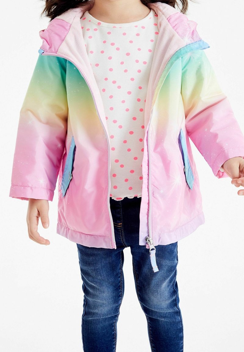 Enfant portant une veste arc-en-ciel pastel par-dessus une chemise blanche à pois roses et un jean bleu, debout les bras légèrement écartés.