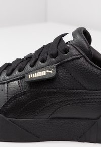 Svart lädersneaker med texturerad yta, med svarta snören, guld logotag och en texturerad svart gummisula.