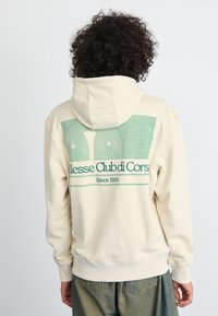 Hanorac de culoare crem, cu un grafic mare verde pe spate, având textul "Alesse Club di Corse Since 1959" într-un design geometric, în dungi.