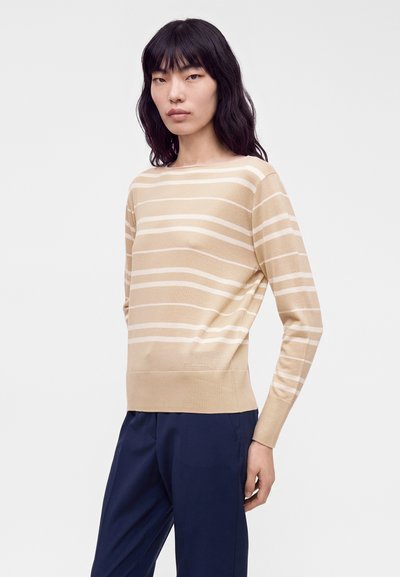 Femme aux longs cheveux noirs portant un pull à manches longues beige et blanc à rayures et un pantalon bleu foncé, debout devant un fond clair uni.