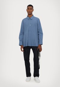 Chemise bleu clair à manches longues avec une partie inférieure plissée et une fermeture à boutons sur le devant, associée à un pantalon en denim noir et des baskets blanches.