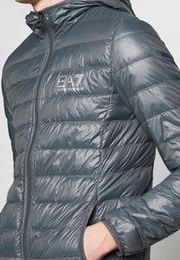 Šedá péřová bunda s kapucí, prošívaným designem, předním zipem a logem EA7 na hrudi. Vyznačuje se strukturovanou tkaninou a přiléhavým střihem.