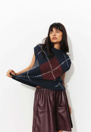 Pull en maille oversized bleu marine et bordeaux en argyle, à manches courtes, porté par-dessus une jupe en cuir bordeaux avec une taille élastique.