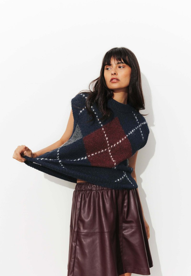 Pull en maille oversized bleu marine et bordeaux en argyle, à manches courtes, porté par-dessus une jupe en cuir bordeaux avec une taille élastique.
