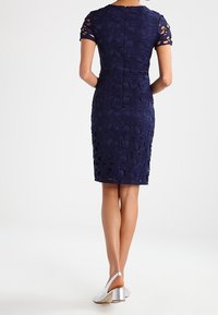 Navy blue fitted dress with a textured floral lace pattern, short sleeves with cut-out details. Worn with silver heeled shoes.