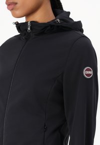 Črna softshell jakna z visokim ovratnikom in nastavljivo kapuco, srebrnim zadrgo in okroglo Colmar oznako na levi rami.