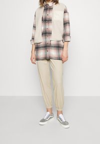 Plaid button-up shirt in black, white, and pink with beige fleece vest. Beige tapered pants and gray sneakers with white laces complete the look.