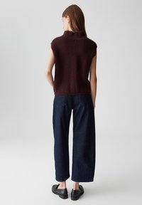 Vrouw staat met de rug naar de camera, gekleed in een mouwloze donkerbruine gebreide top, wijde donkerblauwe jeans en zwarte instappers tegen een effen achtergrond.