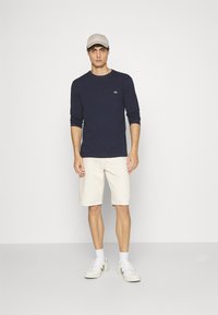 Chemise à manches longues bleu marine avec un logo, short crème et baskets blanches. Le modèle porte une casquette beige clair. Design simple et décontracté.