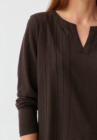 Tunique marron foncé à manches longues, avec des plis verticaux et un col en V. Fabriquée en tissu doux, avec une coupe décontractée et un design minimaliste.