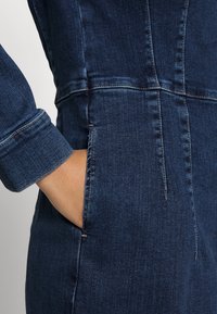 Ręka w kieszeni ciemnoniebieskiego denimu z widocznymi szwami i podwiniętym rękawem.