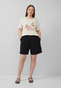 Cremfarbendes T-Shirt mit "Sea La Vie" Grafik, kombiniert mit schwarzen Shorts. Das Outfit hat eine lockere Passform und ein lässiges Design, ergänzt durch minimalistisches Schuhwerk.