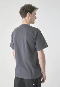 Man with short brown hair wears plain dark gray short-sleeve t-shirt and black pants, shown from the back against a light gray background.
