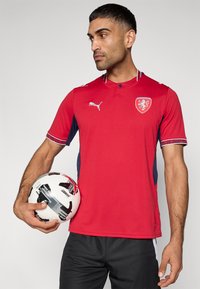 Κόκκινη αθλητική φανέλα με ναυτικό μπλε πλαϊνά πάνελ, κοντά μανίκια, γιακά με κουμπιά και λογότυπο οικόσημο. Μια ασπρόμαυρη ποδοσφαιρική μπάλα κρατείται στο χέρι.