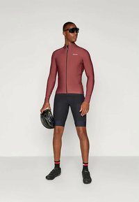 Sportīga riteņbraukšanas apģērba komplekts, kas ietver pieguļošu bordo garām piedurknēm topu un tumši zilus šortus, ar spīdīgu melnu ķiveri rokās un tumšiem saulesbrillēm.