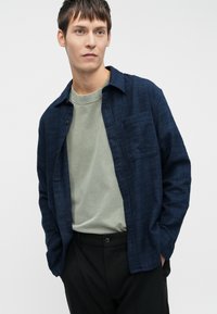 Navyblauw button-up shirt met een gestructureerd patroon, enkele borstzak, gelaagd over een lichtgroene ronde hals t-shirt, gecombineerd met zwarte broek.