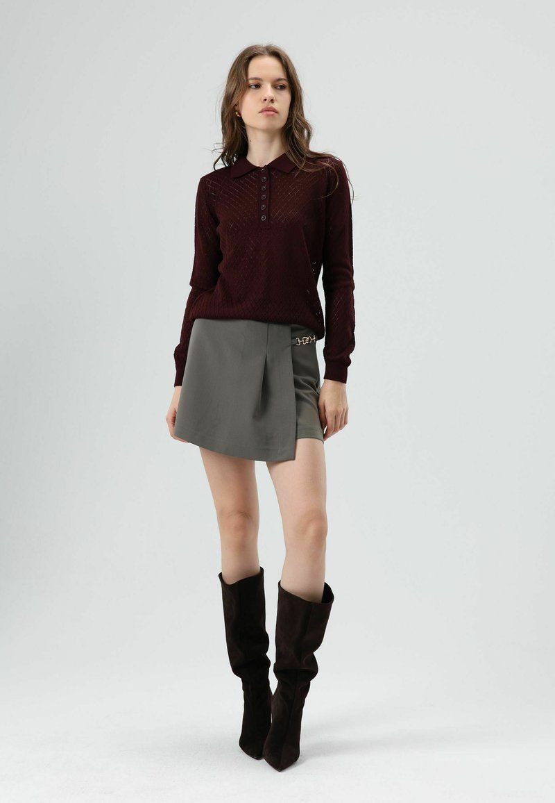 Polo bordeaux à manches longues avec un motif texturé, associé à une jupe-short grise mi-longue avec un pli sur le côté et des accents de quincaillerie argentée. Bottes noires hautes jusqu'aux genoux.