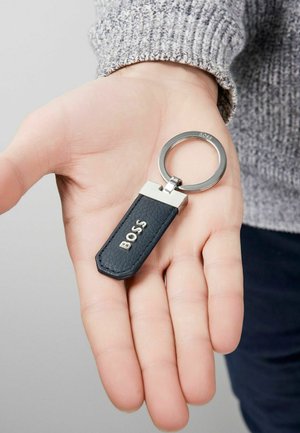 Melnas ādas atslēgu piekariņš ar sudraba aparatūru, ar taisnstūra piekariņu, uz kura ir iepresēts vārds "BOSS". Tekstūrēta virsma, sešstūra forma.