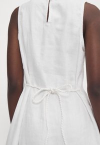 Valkoinen pellavamekko, jossa on avaus selässä ja sidottava vyötärö. Sileä pinta, hihaton, ja flare-helma.