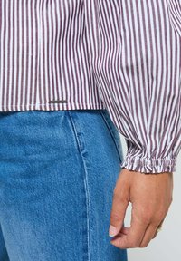 Gros plan sur une main portant une bague en or, une chemise rayée bordeaux et blanche avec des poignets froncés, et un jean en denim bleu.