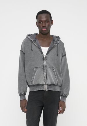 Camisola com fecho de correr cinzenta, capuz com cordão, punhos acanalados e bolsos frontais. Apresenta costura contrastante e corte descontraído.