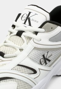 Sneaker značky Calvin Klein Jeans s bielo-šedou sieťovinou, čiernymi akcentmi a textúrovanou gumovou podrážkou s detailmi loga.