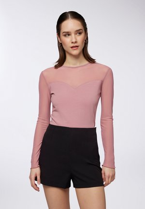 Maglietta a maniche lunghe di colore rosa chiaro con pannello superiore in rete trasparente e scollatura tondeggiante, abbinata a pantaloni shorts neri a vita alta. Tessuto dal tatto liscio.