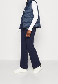 Granatowa pikowana kamizelka puchowa z wysokim kołnierzem, zestawiona z białą bluzką z długim rękawem oraz granatowymi spodniami o szerokich nogawkach, uzupełniona białymi trampkami.