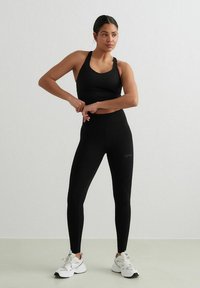 Crni atletski crop top s dizajnom na leđima u obliku trkača u kombinaciji s crnim tajicama visokog struka. Obje su napravljene od glatkog, rastezljivog materijala. Bijele teniske.